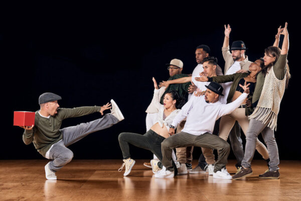 A dancer squats holding a red box in front of a group of dancers who are in different poses seeming to recoil.