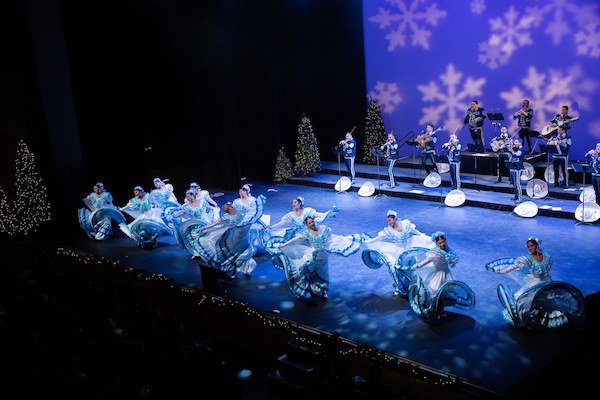 The stage is filled with mariachi musicians and Mexican folkloric dancers in blue swirling skirts.