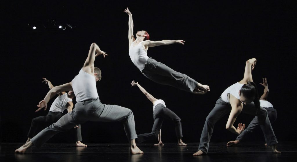Dancers in grey leggings and white shirts jump and pose