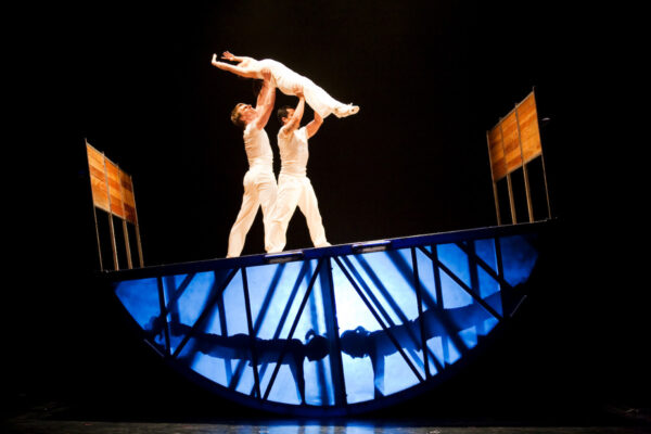 Two dancers on top of a structure hold a third dancer in the air