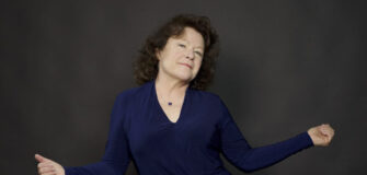 Photo of Beth Ruscio, a Caucasian woman, looking at the camera with her arms spread out, in front of a dark background.