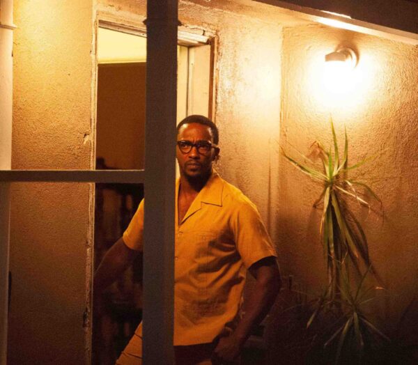 Image of a Black male, the actor Anthony Mackie, standing outside an open door under a yellow tinted light at night, staring off to the left of the frame. He is wearing glasses and a short sleeve button down shirt.