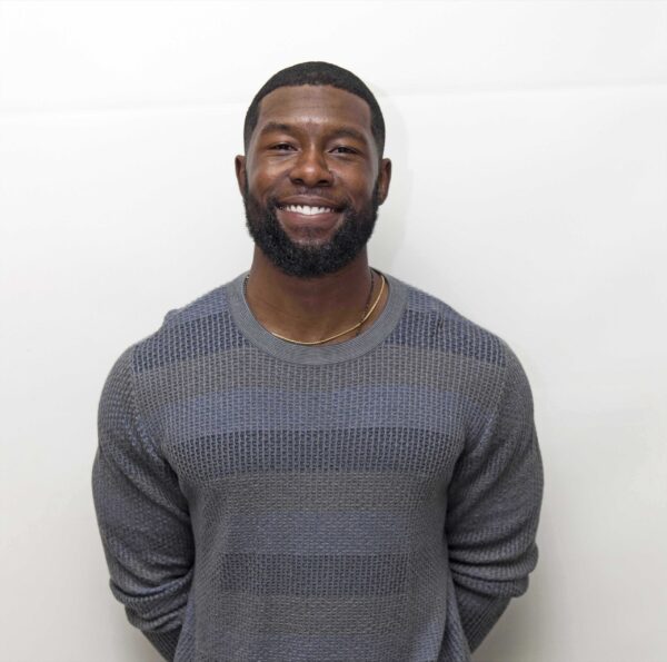 Image of Black American actor Trevante Rhodes standing with his arms behind him in front of a white wall. He is wearing a striped blue and gray sweater. He is smiling into the camera.