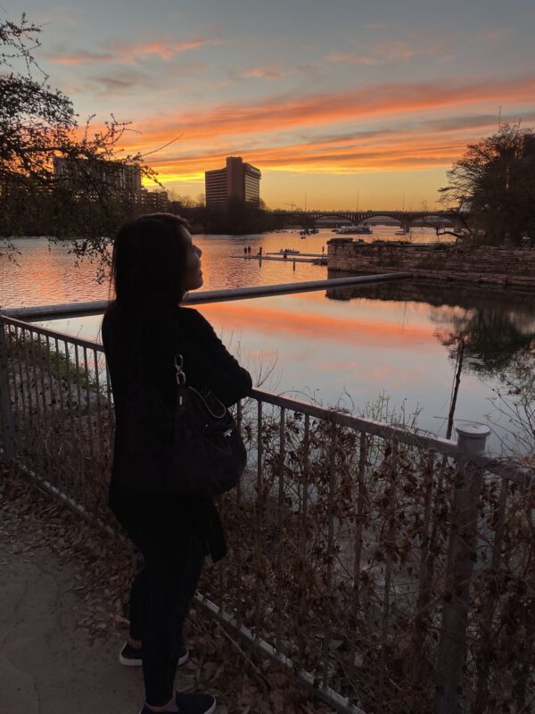 An image of an Asian woman standing at a railing by a body of water, staring right of frame at the orange, gray, yellow sunset.