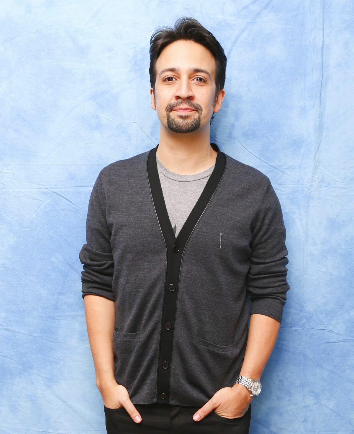 In this picture, the genius award winning Lin Manuel-Miranda stands relaxed, his hands in his pockets, wearing a cardigan in front of a blue backdrop.