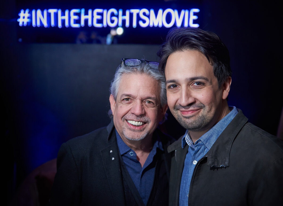 A really sweet photo of Lin-Manuel Miranda with his father Luis, who looks so proud of his son. They're even wearing matching outfits, dark jacket and blue button down shirt, with the hashtag for In The Heights Movie in neon behind them.