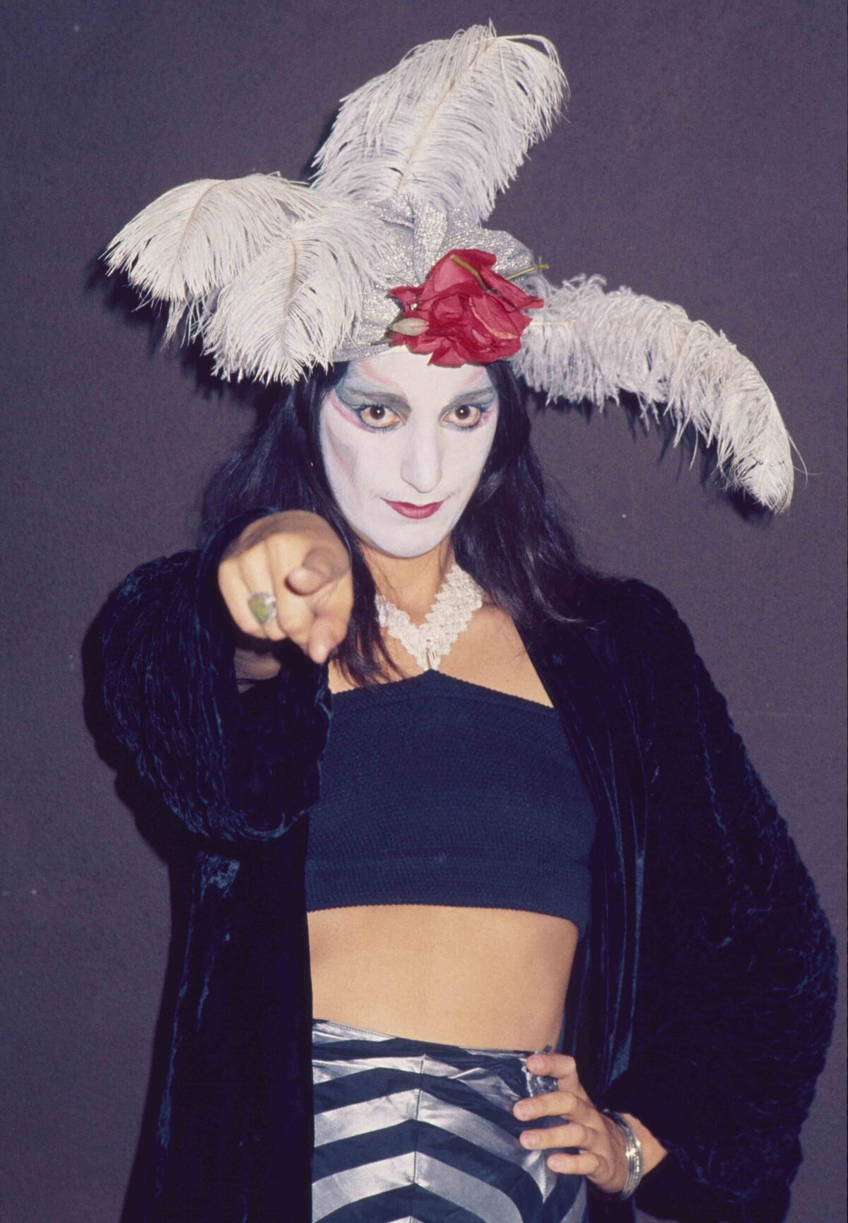 A person in white mime face paint, with three matching white feathers on her head, points at the camera.