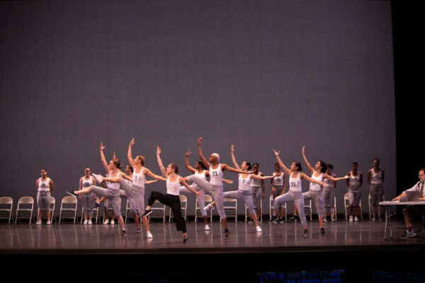 A large group of people in sweatpants and white tops dance together on a large stage.
