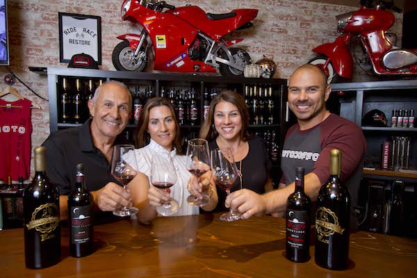 A smiling foursome, the Doffo family, each holding up a glass of red wine to toast. Behind them are bottles and bottles of wine on a shelf, which, by the way, happens to have a hot red motorcycle (and a red scooter) sitting atop.
