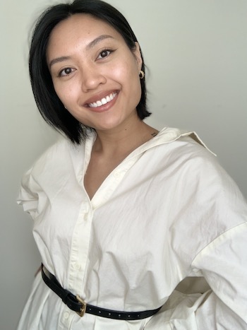 Kendra Quadra smiles, her head slightly tilted to one side, her long white button up shirt tied at the waist with a thin black belt.