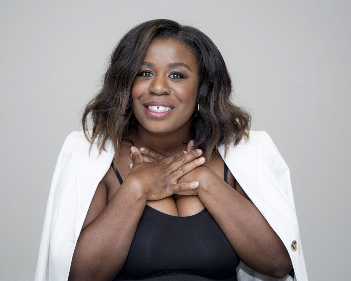 Uzo Aduba with her hands crossed at her chest, a big smile showing her trademark gap. Her hair is straight and a white jacked hangs on her shoulders.