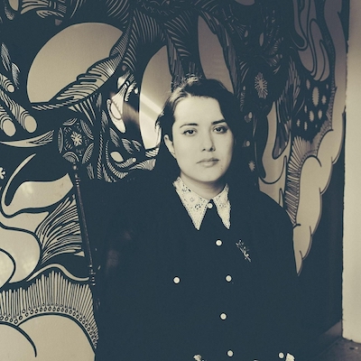 A black and white image of a person, Viva Padilla, standing in front of a mural.