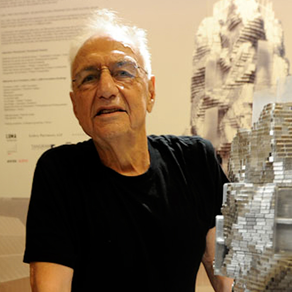 Frank Gehry, his white hair striking in contrast to his black t-shirt, stands next to a model of one of his designs.