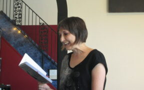 Poet Juliet Cook is Caucasian with brown hair; she's standing up eading from a book. She's smiling.