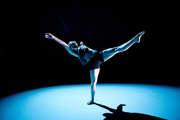 A female dancer in a blue spotlight stands on one leg and extends the other in the air