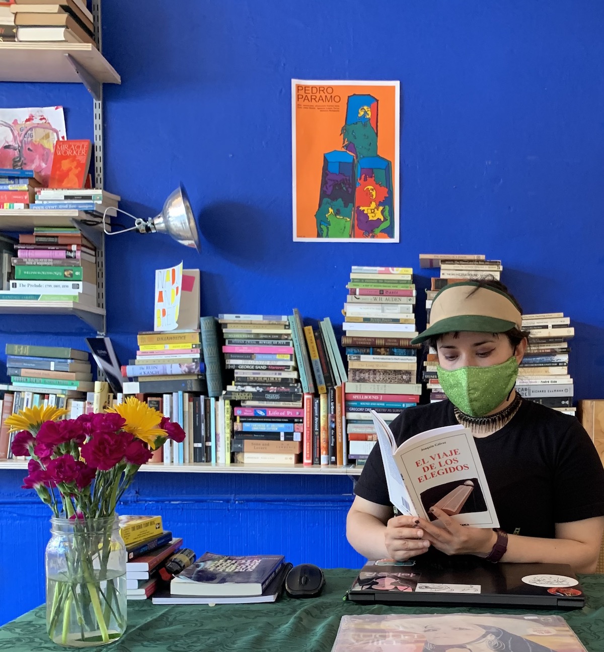 A person in a mask (Viva Padilla, founder of Re/Arte) reads a book called El Viaje De Los Elegidos inside a blue walled room full of stacked books.