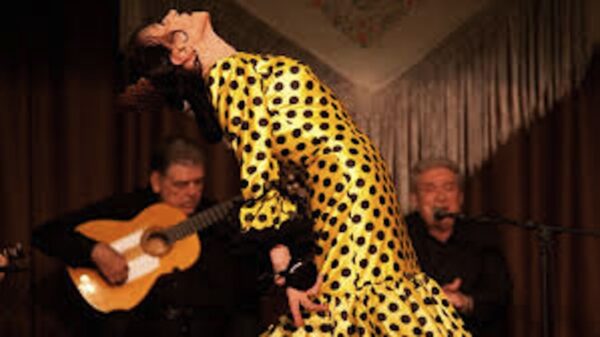 A flamenco dancer in a yellow dress with black polka dots bends back toward the guitarist playing behind her.