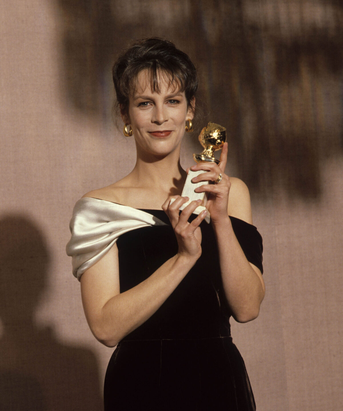 Jamie Lee Curtis, wearing a black and gold dress, holds up her trophy with a smile on her face.