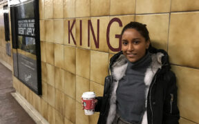 A young Black woman leans against a wall, the word KING on the tiles. She is holding a to go cup of coffee.