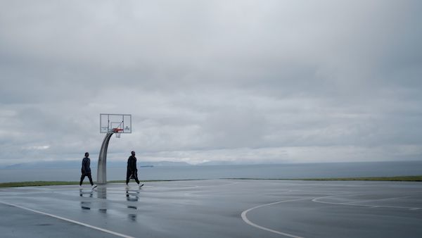 Two people walk by on an empty basketball court under a cloudy sky