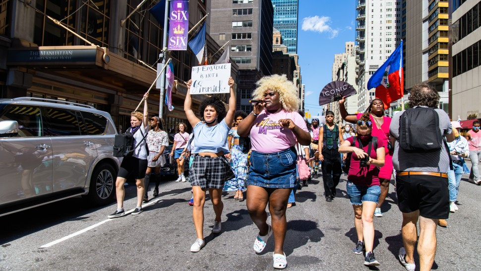 Two Black trans women lead a march with one holding a sign that reads "My existence is not a gimmick" while the other holds a microphone.