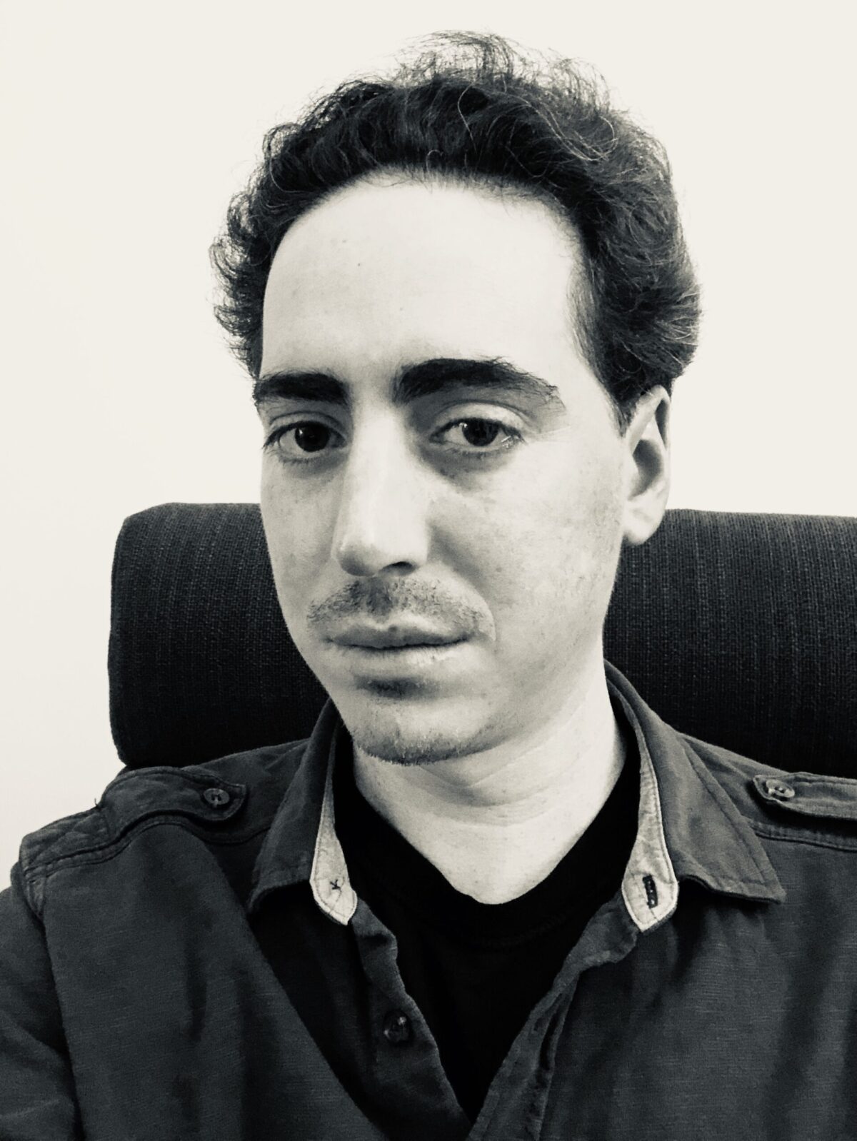 A black-and-white photo of poet Mark Danowsky sitting in a chair and looking at the camera. He has a five o'clock shadow on his upper lip, lazy eyes, and a black shirt underneath a jacket. 