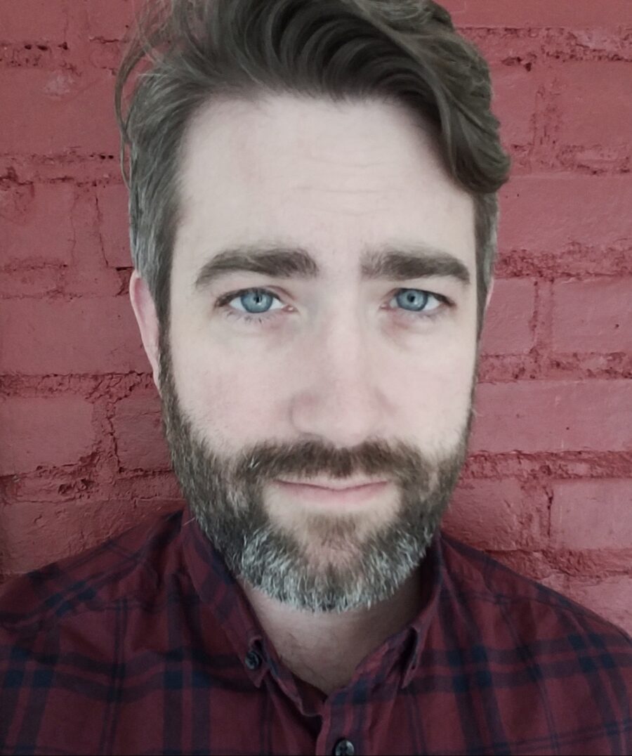 Headshot of poet James Duncan. He's in front of a red brick wall; full grayish beard, with blue eyes staring directly at camera.
