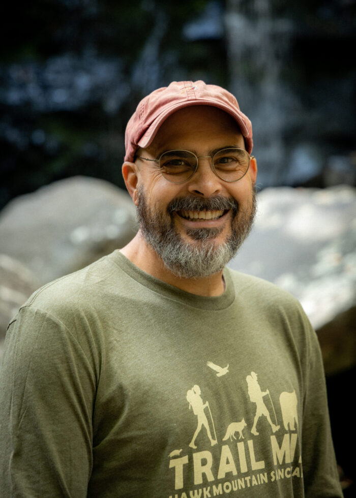 Photo of poet Grant Clauser. He's wearing a green t-shirt with the word "Trail" on it, turning sideway and smiling at the camera. He wears a light pink hat, glasses, and full beard. In the background is the great outdoors. 