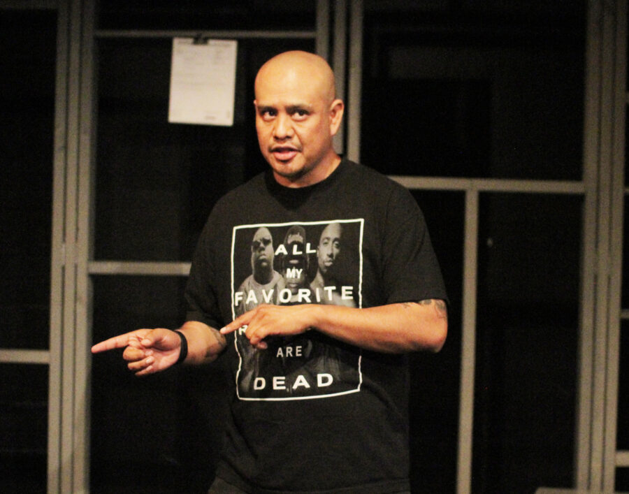 Actor Alex Alpharaoh on stage in his show WET, in a black t-shirt pointing at something off stage to this right.