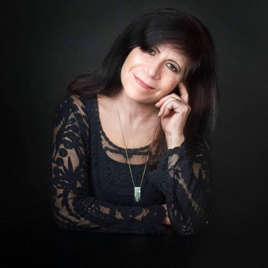 Photo of poet Cynthia Atkins. Black background. Cynthia has long dark hair. She tilts her head to the left. Her left hand gently holding her head. She has on a long silver pendant.