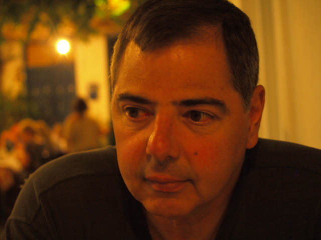 A closeup photo of poet John Tripoulas. He is either outside or in a restaurant, and it's in the evening. He might be sitting at a restaurant. He looks to his right. he has a mole near his left nose, on his left cheek.