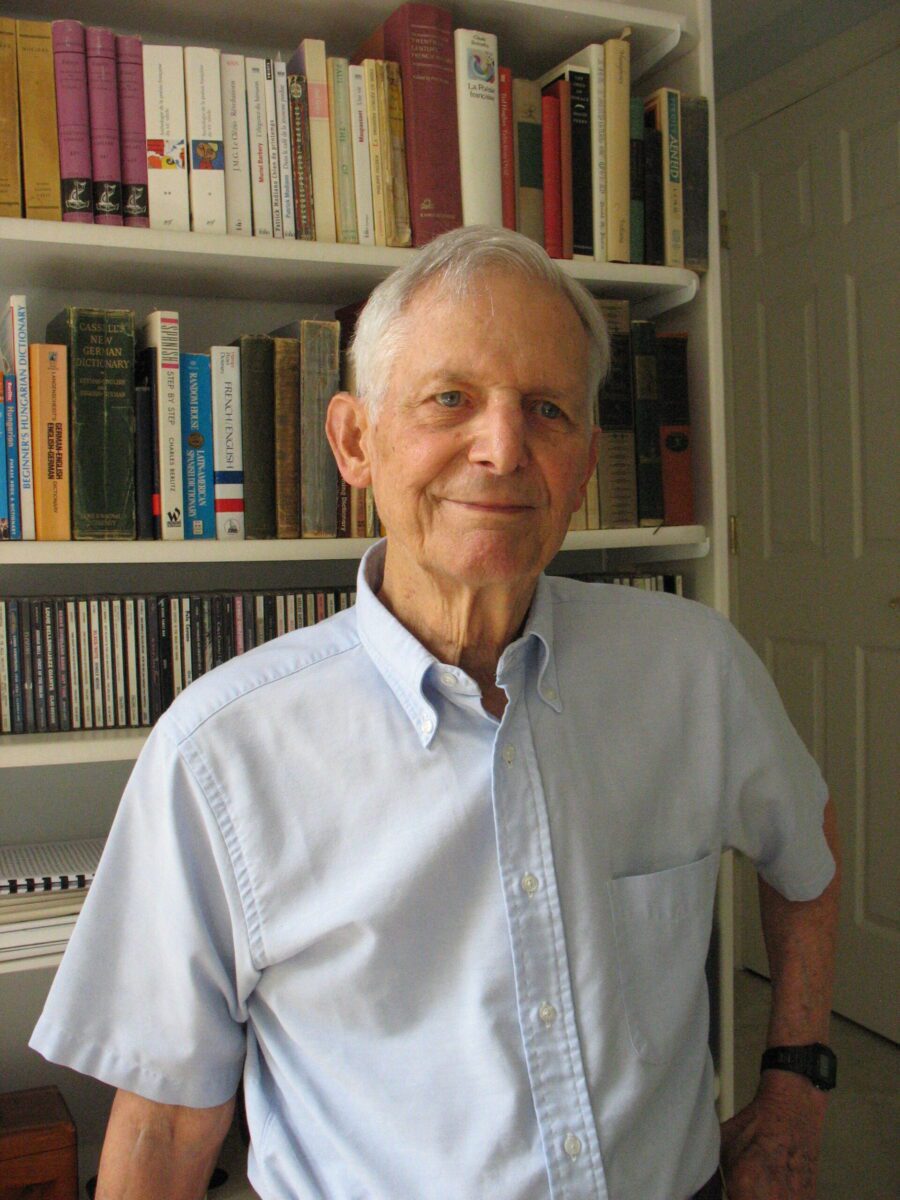 Photo of poet and professor Leonard Trawick. He is in a light blue shirt, standing in front of his book case, hair is white, parted to his his right. His left hand is on his waist. He is smiling with mouth closed.