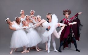 Dancers from Swan Lake hold one dancer while a dancer in black holds that dancer's leghree women ballet dancers are in different upright poses and a man is entering the area as if running.