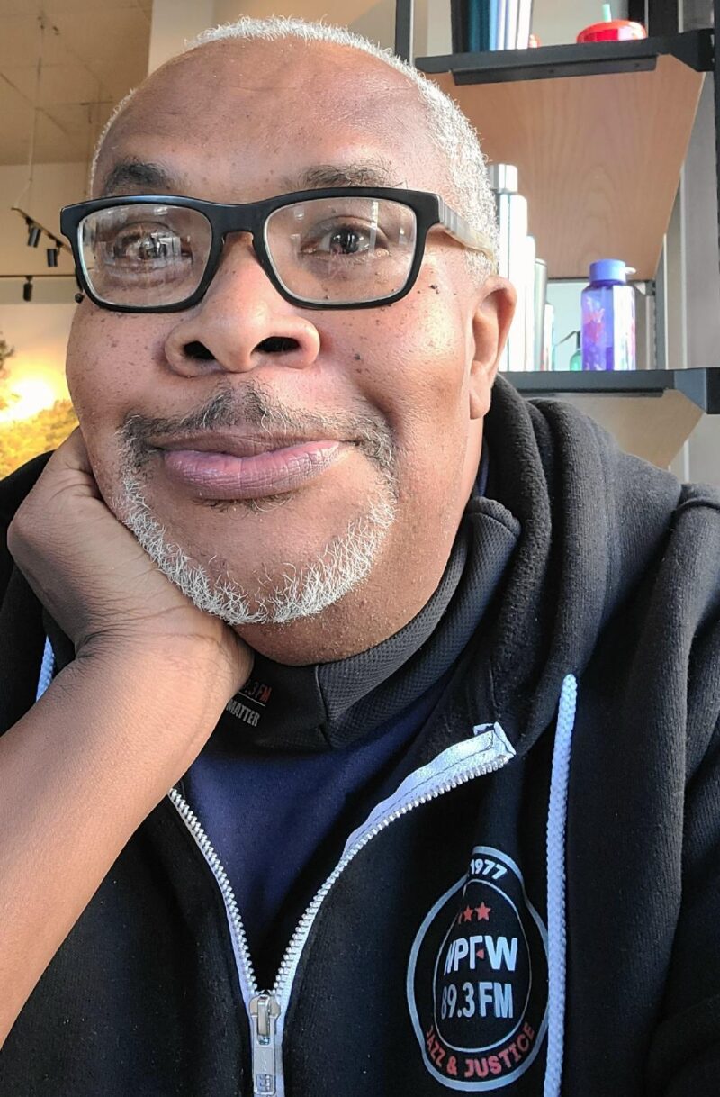 Close-up photo of poet Reuben Jackson. He's wearing a hoodie with the radio station that he works for. Reuben wears black-rimmed glasses, his head resting on his hand, and smiling hopefully at the camera.