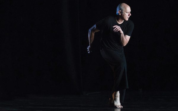 A bald man in black stands on one foot