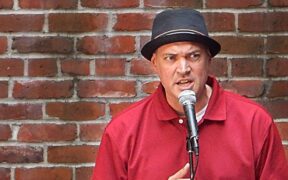 Mike Sonksen, in a red shirt and a hat, performs at the mic