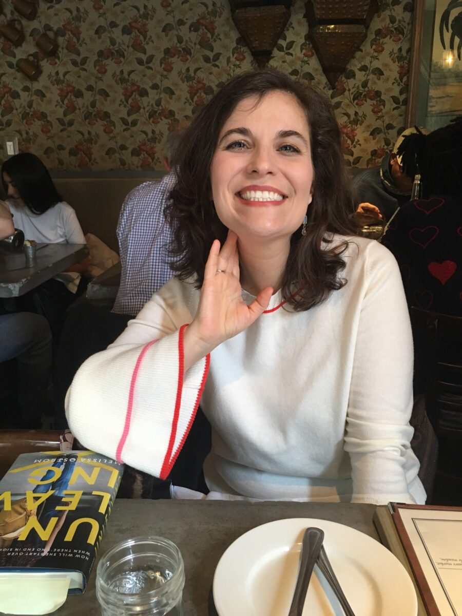 Photo of author Melissa Ostrom. She is sitting at a table inside a crowded restaurant; this is most likely before the pandemic. She has on a white dress and is smiling at the camera. On the table is a copy of her second novel, The Unleaving.