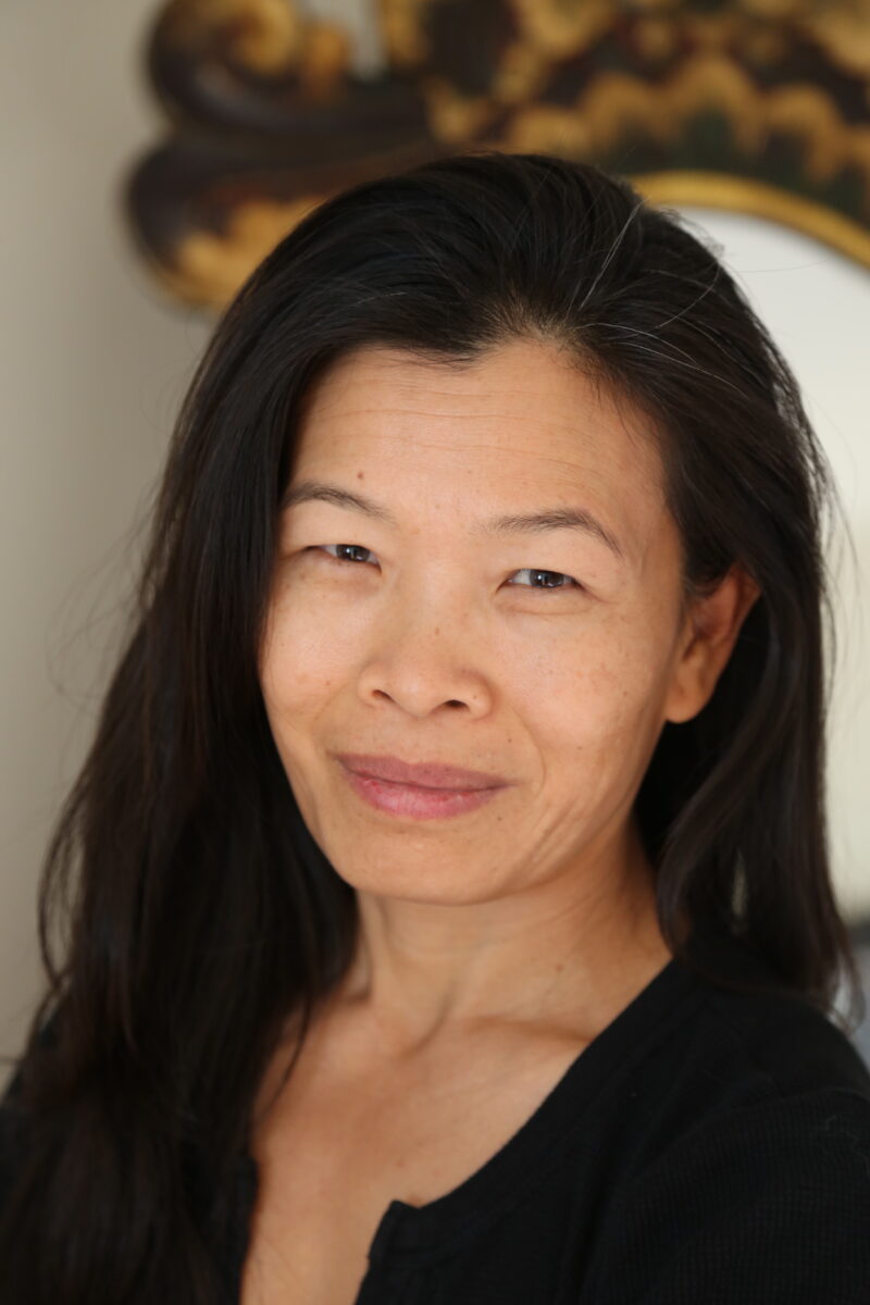Color photo of Cambodian-American poet and yoga instructor. She has long black hair, starring and smiling sideway at the camera. Her skin is radiant without makeup. 
