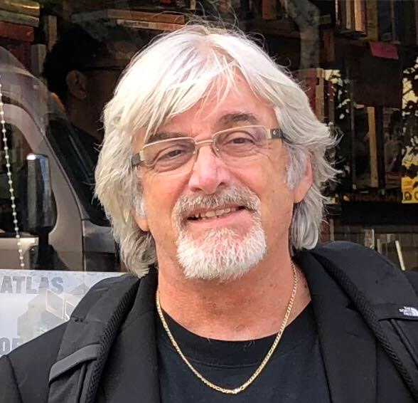 Photograph of Al Ortolani. Al has white hair and bear, with glasses, and smiling. He has on a black jacket, black t-shirt, and a necklace around his neck. He looks at the camera, smiling somewhat, and looks casual, easy going.