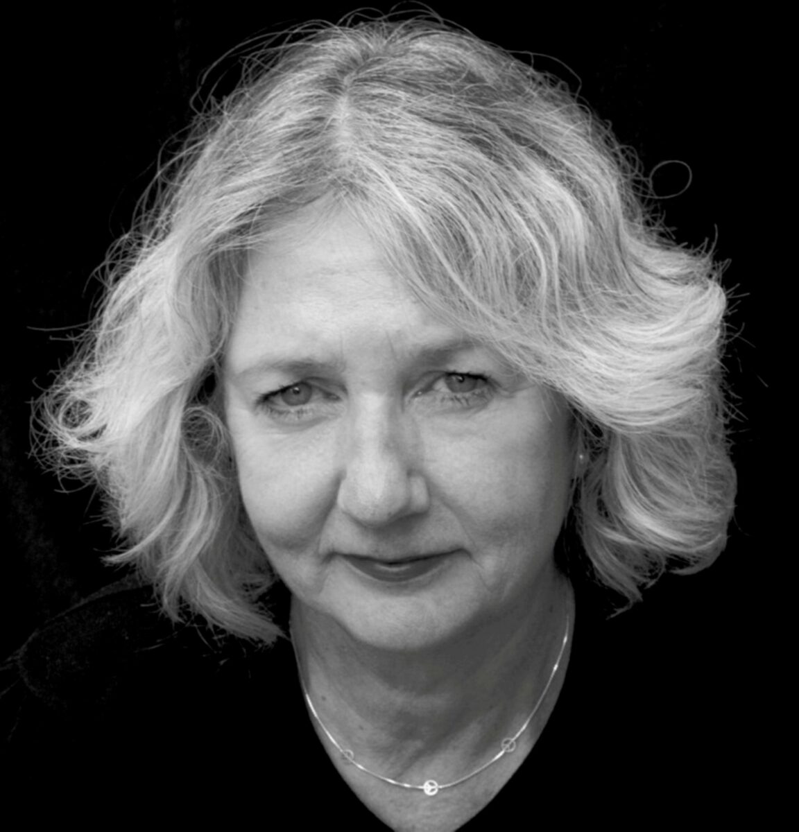 A black-and-white photo of poet Cora Siré. In a black background, black shirt, she has blondish/grey hair, wearing necklace and possibly red lipstick.She stares directly at the camera.
