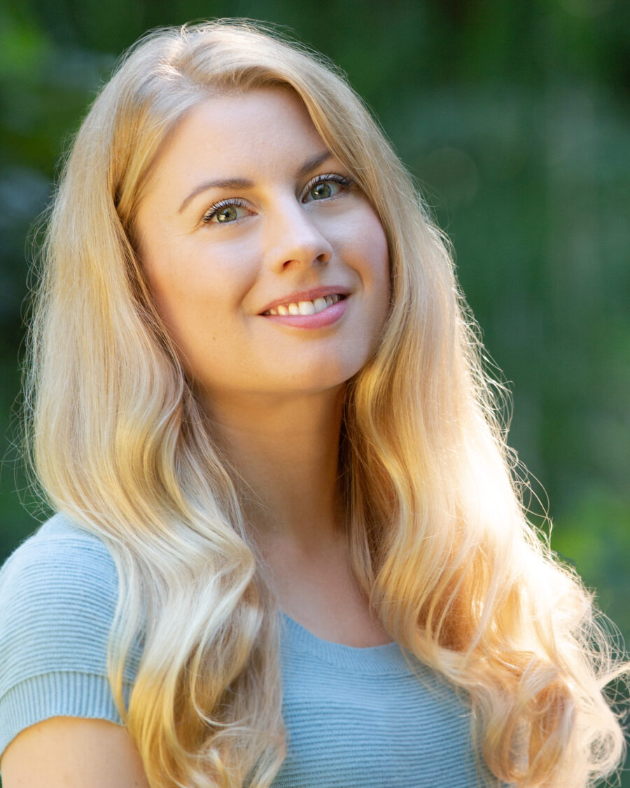 Headshot of Canadian poet Emily Osborne. She has long blonde hair; her head tilts upward as she smiles at the camera. The greenery in the background is not focused.
