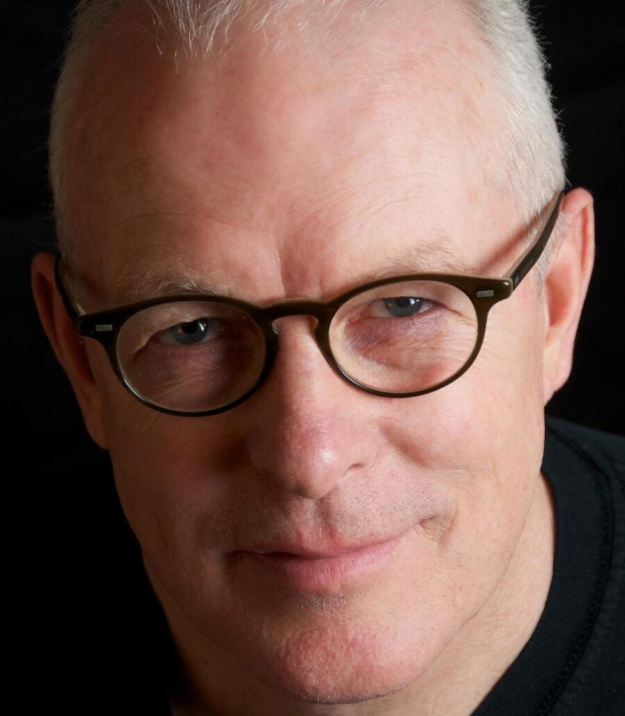 Photo of poet Kevin Carey. Very close-up shot of his face. He wears black-rimmed glasses and short grey hair. Background is black.