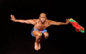 A bare chested man leaps with his arms extended