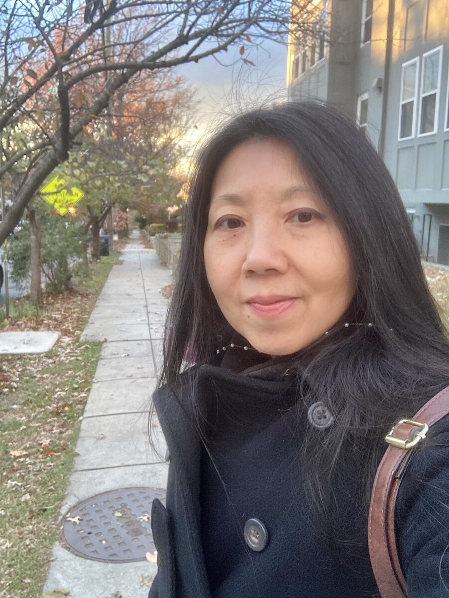 Photograph of Hmong-American-French poet Pacyinz Lyfoung. She is on the sidewalk, possibly in fall weather, starring at the camera in the selfie. She has on a black coat.