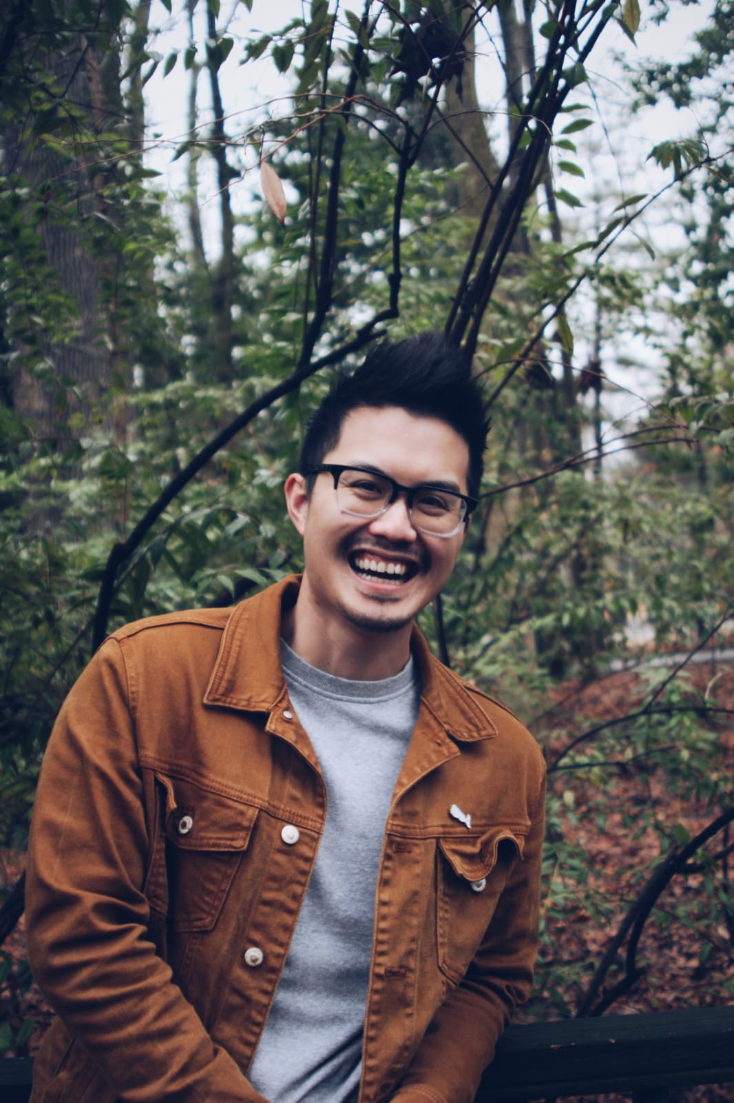 Photo of poet Joshua Nguyen. He is outside, possibly in the park. He wears glasses and has a big smile.