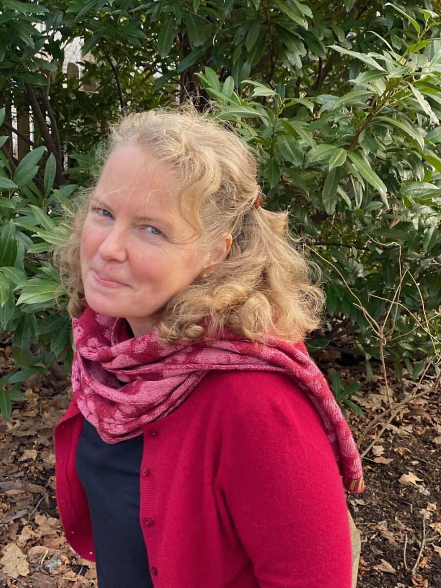 color photo of poet Kasey Jueds. She's outside in red shirt, pink scarf around her neck, sitting sideway and smiling at the camera. Bushes in the background.