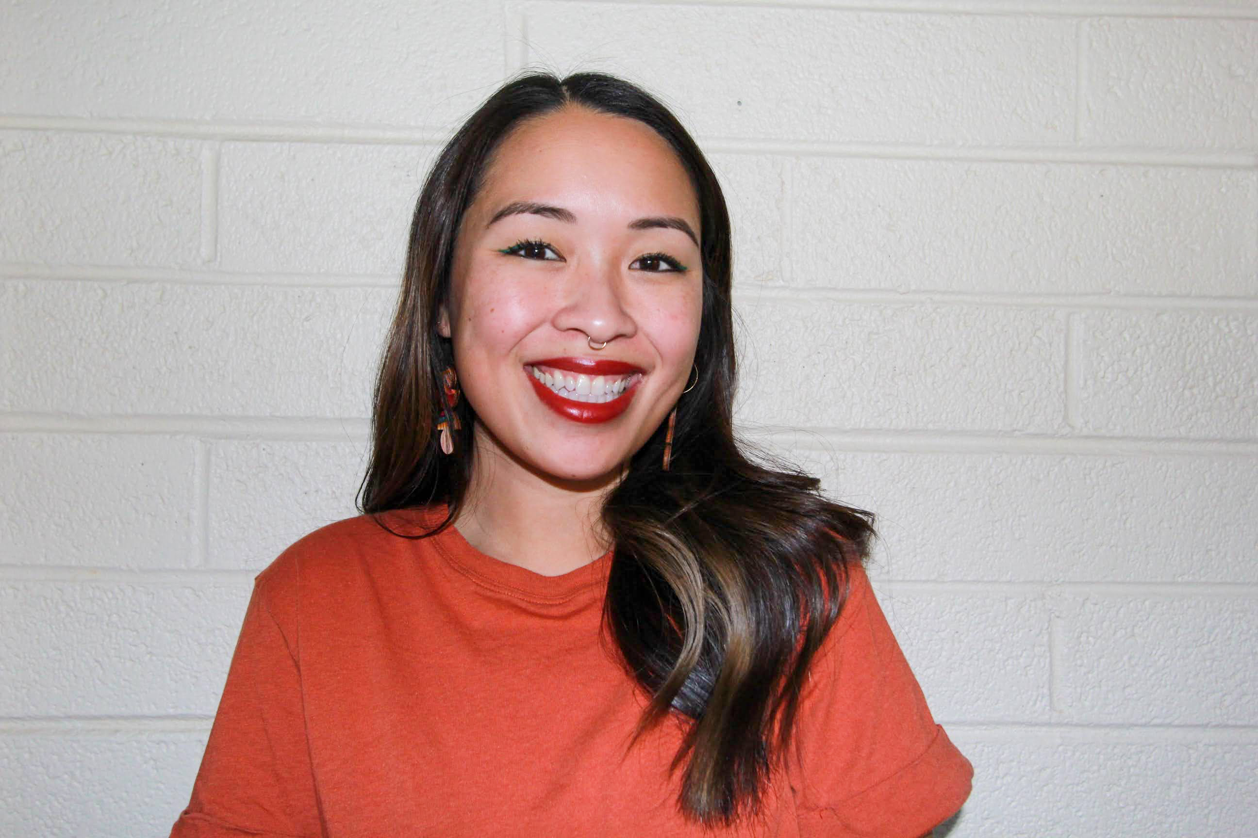 Photo of poet Susan Nguyen. She is in a red shirt; her long hair (with some highlights) rests on her left shoulder. She sports a nose right and she smiles brightly at the camera.