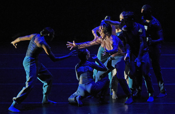 A groups dancers in blue reach toward a male dancer