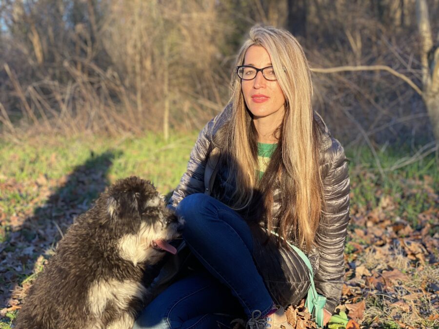 Photo of poet Alina Stefanescu. She is sitting outside with her dog next to her. Sun on her face and long blonde hair, with glasses and sweater. 