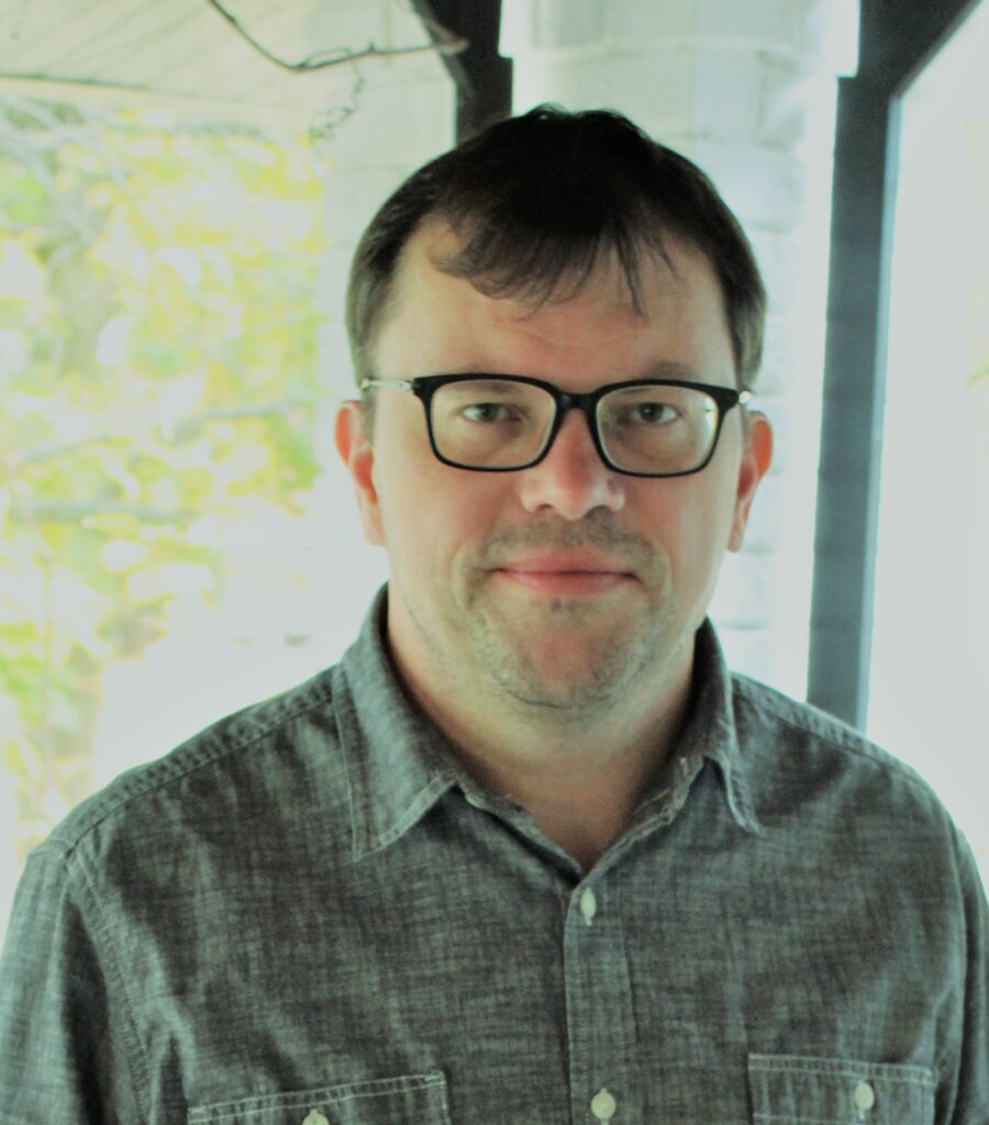 Color photo of poet and professor Chris Haven. He's on black-rimmed glasses, short hair, and a bluish grey collar shirt. He's looking directly at the camera.